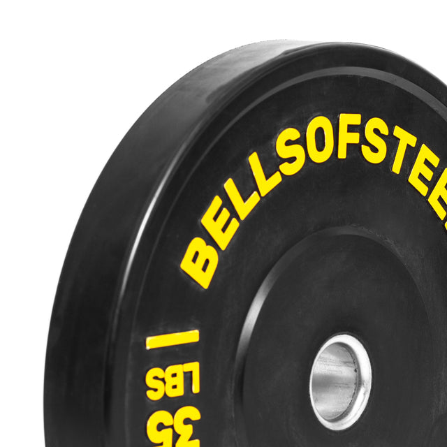 A close-up of a Bells of Steel Dead Bounce Conflict Bumper Plate Set in black virgin rubber, with yellow branding and "35 LBS" text, plus a durable stainless steel insert around the center hole.