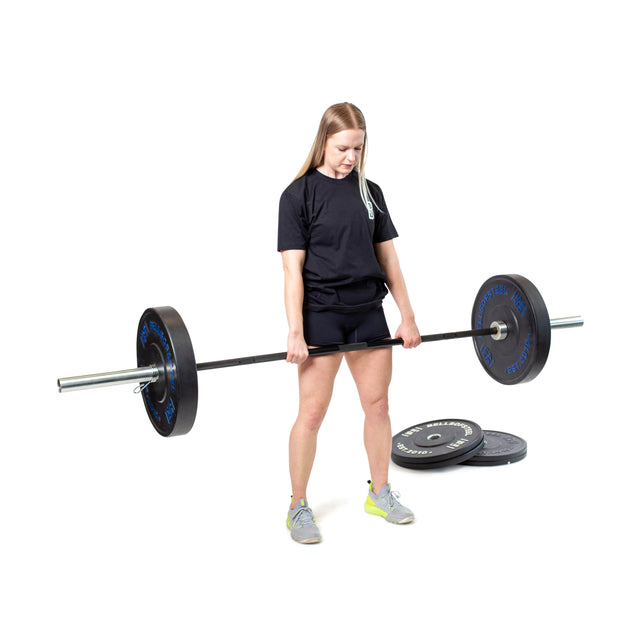 A person in a black t-shirt and shorts lifts a loaded barbell with Bells of Steel Dead Bounce Conflict Bumper Plate Sets, made from virgin rubber with stainless steel inserts. Extra plates are stacked nearby against a white backdrop.