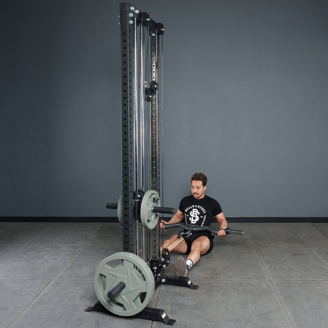 A person sits on a gym floor, holding the Bells of Steel Fat Bar - Olympic Curl Bar Cable Attachment on a tall cable machine with weight plates, preparing for a seated row. The background has plain gray walls and flooring.