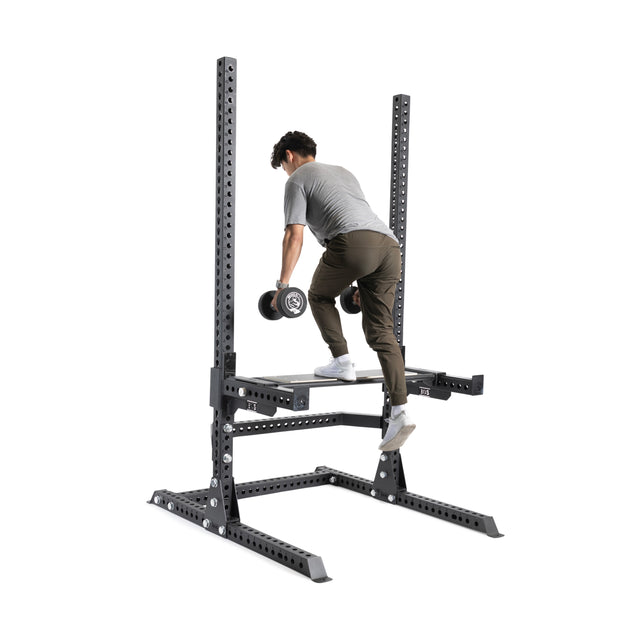 Wearing athletic gear, a person uses a dumbbell to perform step-ups on the Bells of Steel Utility Seat for Hydra/Manticore, attached to a sturdy 3"x3" rack with adjustable platforms.