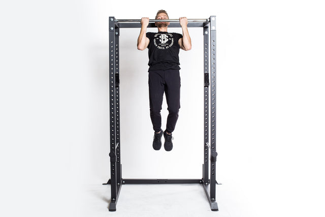 Wearing black athletic clothing, a person does a pull-up on the Bells of Steel Residential Power Rack (2.3" x 2.3", ⅝" holes) with j-cups, set against a plain white background.