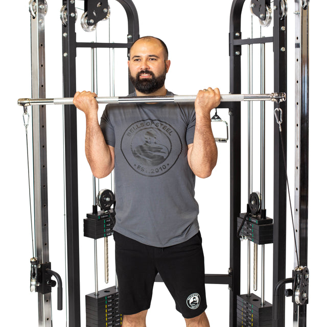 A bearded man in a gray T-shirt and black shorts is using the Bells of Steel Double Hook Straight Bar Cable Attachment, standing between the cable machine’s frames and holding the bar at chest height for his workout.
