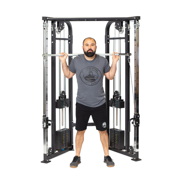 A man stands at a cable machine, holding the Bells of Steel Double Hook Straight Bar Cable Attachment across his shoulders and upper back. He wears a gray t-shirt, black shorts, and black sneakers, facing forward with a neutral expression.