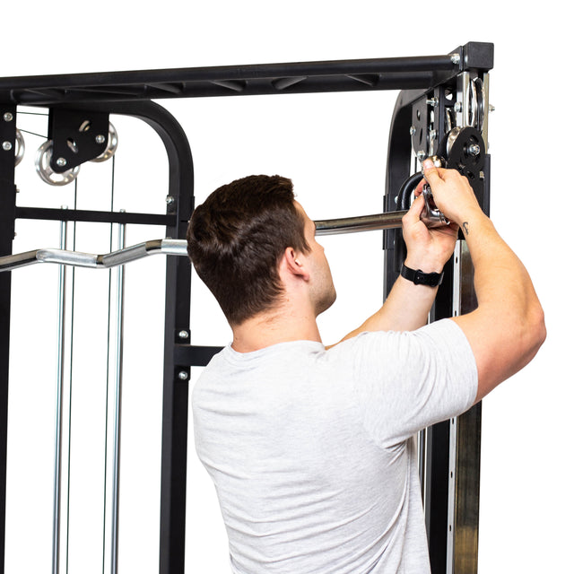 A man in a light gray T-shirt adjusts a cable machine with the Bells of Steel Double Hook EZ Curl Bar Cable Attachment, perfect for joint-friendly barbell exercises, against a white background.