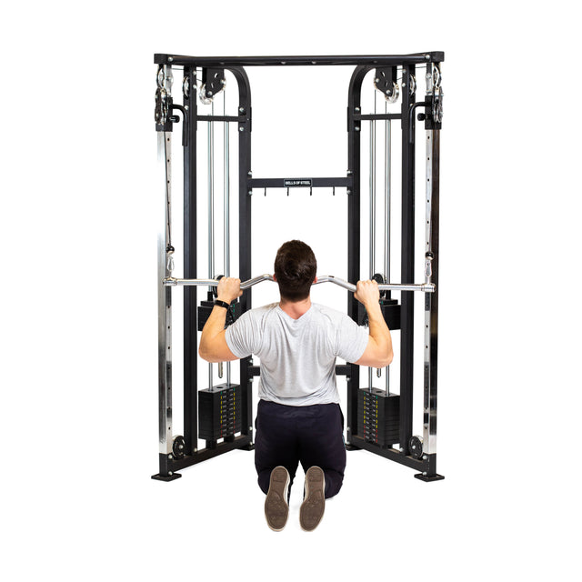 A person in a grey t-shirt and dark pants kneels, using the Bells of Steel Double Hook EZ Curl Bar Cable Attachment with both hands during a strength training exercise on a cable machine.