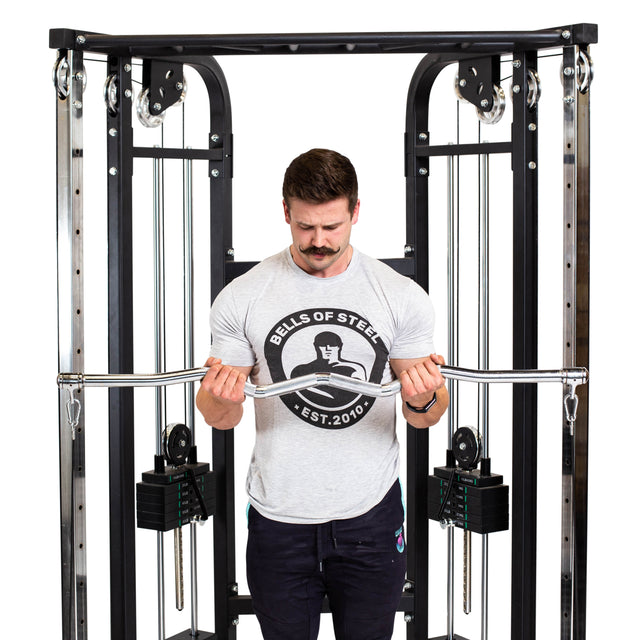A man in a Bells of Steel t-shirt does a bicep curl with the Double Hook EZ Curl Bar Cable Attachment, positioned centrally for joint-friendly barbell exercises.