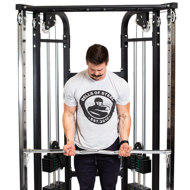 A mustached man in a Bells of Steel t-shirt uses the Double Hook EZ Curl Bar Cable Attachment for joint-friendly barbell curls on a cable machine, set against a white background.