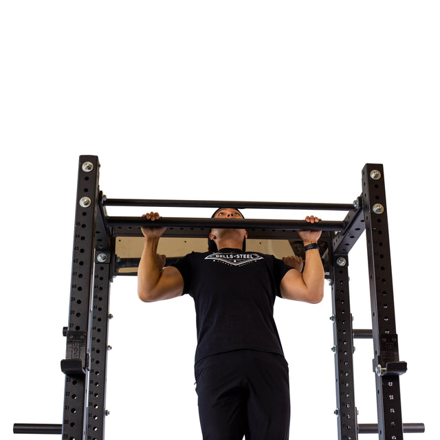 A person wearing black clothes is doing a pull-up on the Bells of Steel Fat/Skinny Pull Up Bar, which is attached to a sturdy power rack. The background is entirely white.