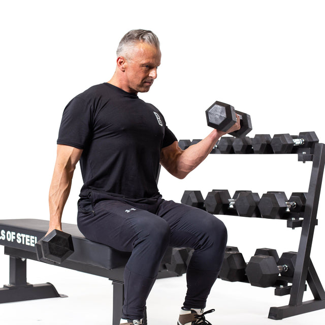 A man in athletic wear sits on a bench, curling Bells of Steel Ergo Rubber Hex Dumbbell Sets. Next to him, a rack of these durable dumbbells stands against a white background.