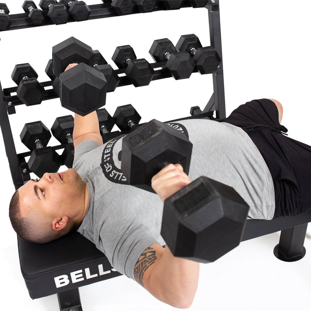 A person lies on a bench holding two Bells of Steel Ergo Rubber Hex Dumbbells in a chest press position, with a rack of Ergo Rubber Hex Dumbbell Sets visible behind them. The person wears a gray t-shirt and black shorts.