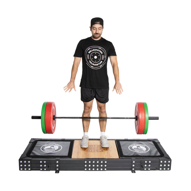 A man in athletic wear and a hat stands on a Bells of Steel Weightlifting Platform with black rubber mats, holding a barbell with red and green plates, focused intently on his lift.