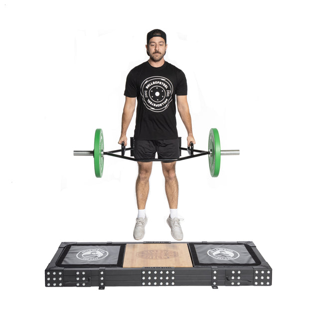 A man stands on a Bells of Steel Weightlifting Platform, lifting a trap bar with green weights. He's dressed in a black cap, logoed T-shirt, black shorts, and white sneakers against a white backdrop. The platform prominently features its logo.