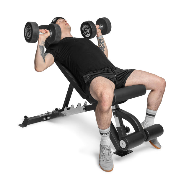 Sporting tattoos, a cap, gray sneakers, and white socks, a person in a black shirt and shorts performs a dumbbell bench press on the "Bells of Steel" Flat / Incline / Decline Weight Bench - Commercial. The plain white background enhances their focused muscle-targeting effort.