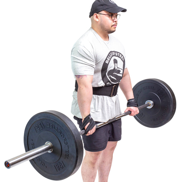 Wearing a Bells of Steel t-shirt and using Classic Figure 8 Lifting Straps by Bells of Steel, a person in a cap, glasses, gloves, and shorts faces slightly right while holding a loaded barbell against a white background.