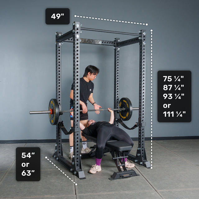 A person spots someone else performing a bench press inside a Bells of Steel Manticore Flat Foot Power Rack Builder (3" x 3", 1" Holes); the rack’s dimensions are labeled as 49" wide, either 54" or 63" deep, and available heights of 75¼", 87¼", 93¼", or 111¼".