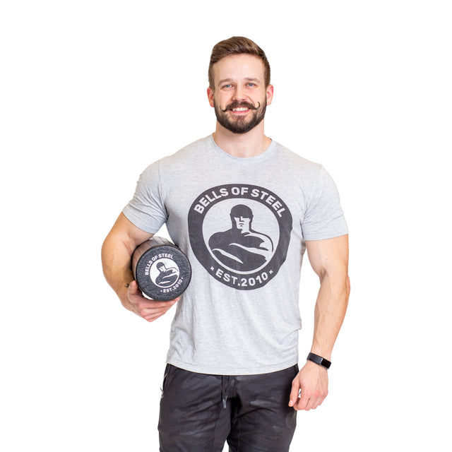 A smiling man with a beard holds a Bells of Steel Foam Roller, perfect for self-myofascial release, and wears a grey “Bells of Steel Est. 2010” t-shirt and black pants, standing against a white background.