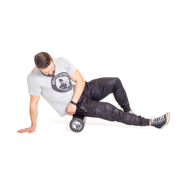 A man in a gray t-shirt and black pants uses the Bells of Steel Foam Roller under his right thigh, supporting himself with his left hand on the floor as he performs a self-myofascial release exercise.