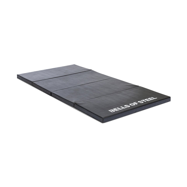 The Bells of Steel Four Fold Mat, a black non-slip workout mat with four folding sections and "BELLS OF STEEL" in white lettering on one end, lies flat on a white background.