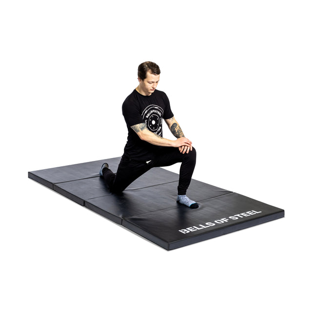 A person in black athletic wear kneels on a Four Fold Mat by Bells of Steel, performing a lunge stretch against a white background.