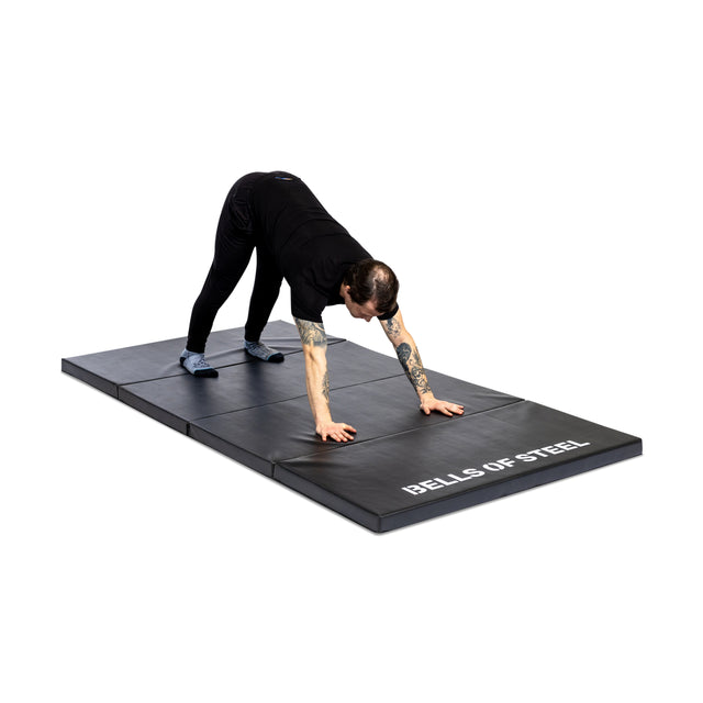 A person in black clothing stretches on a Bells of Steel Four Fold Mat, with hands and feet on the mat and hips raised against a plain white background.