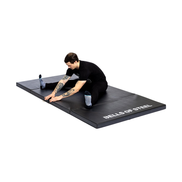 A person in black workout clothes stretches on a large, non-slip Four Fold Mat from Bells of Steel, set against a white background.