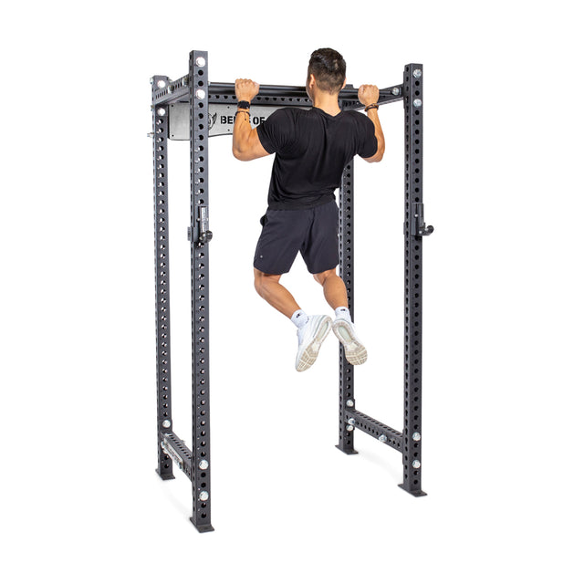 A person in a black shirt and shorts performs a pull-up on the Bells of Steel Fat/Skinny Pull Up Bar, which is built into a sturdy power rack with multiple adjustment holes.