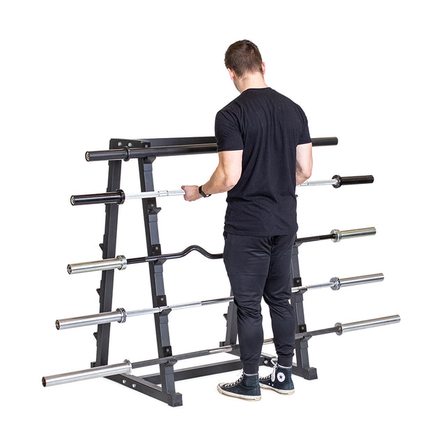 A man in black athletic clothing stands facing the Bells of Steel Fixed Barbell Rack, which holds multiple barbells of various types and sizes, against a white background.