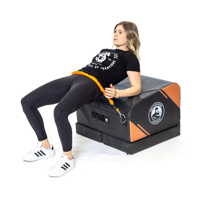 A woman in athletic clothing performs hip thrusts at home using the Bells of Steel Soft Glute Bench and a resistance band, with her shoulders on the bench and feet flat for an effective strength-building workout.
