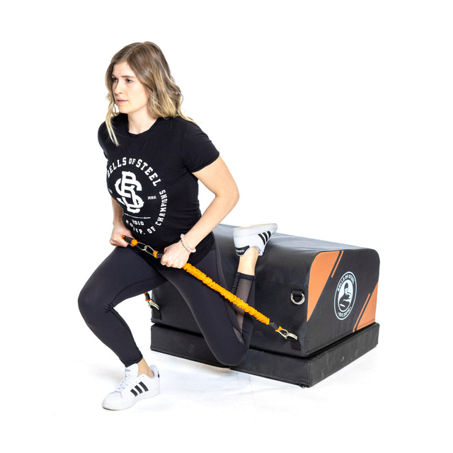 A woman does a Bulgarian split squat at home, using the Bells of Steel Soft Glute Bench as a padded platform and holding resistance bands, against a white background.