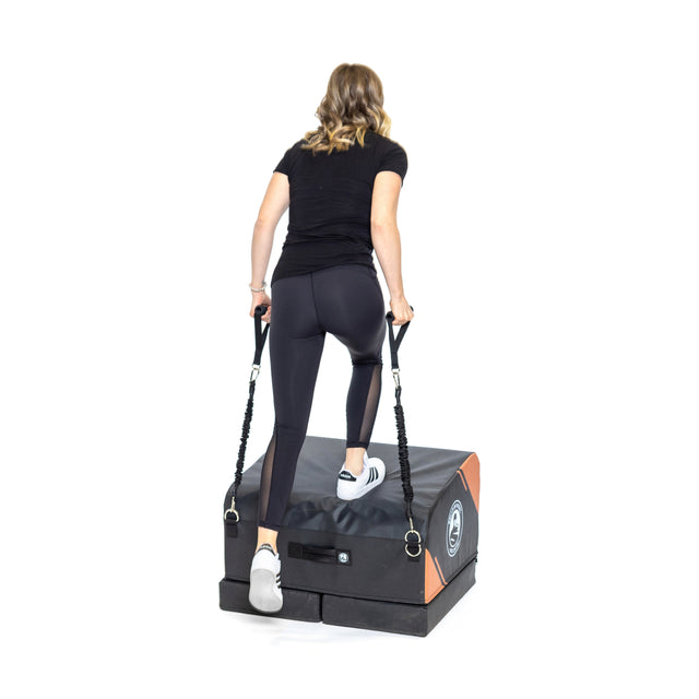 A woman in athletic wear stands on a Bells of Steel Soft Glute Bench, holding resistance bands, viewed from behind against a white background.
