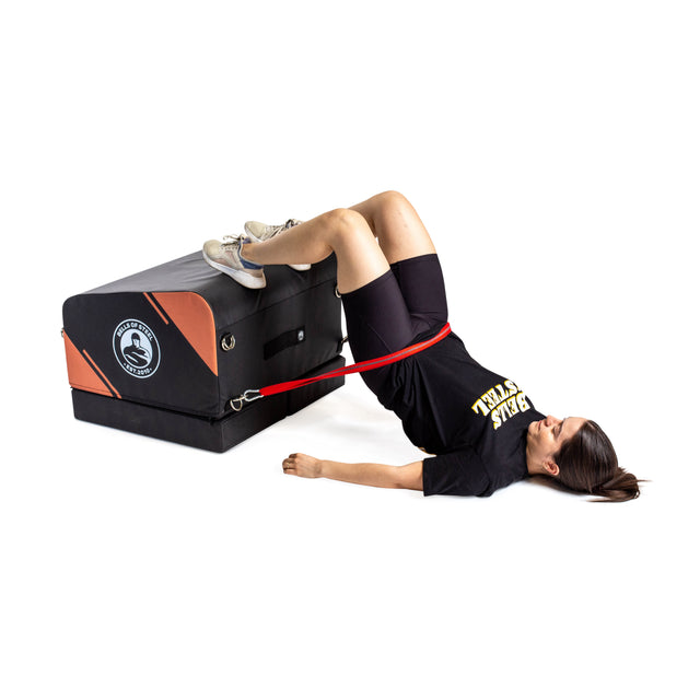 A woman in athletic wear uses the Bells of Steel Soft Glute Bench to perform glute bridges, her feet elevated on the bench and a red resistance band around her hips.