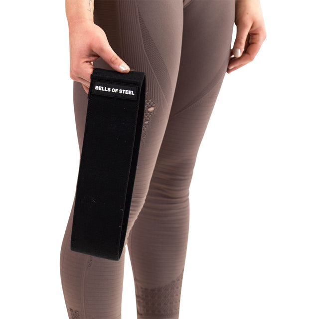 A person in taupe leggings holds a black Bells of Steel Glute Loop resistance band in their left hand, ideal for booty workouts, against a plain white background.