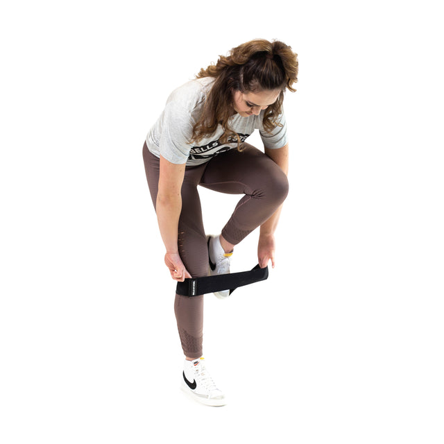 A woman in a gray T-shirt, brown leggings, and white sneakers bends over to put on the Bells of Steel Glute Loop, a black non-slip resistance band, around her legs for a booty workout against a white background.