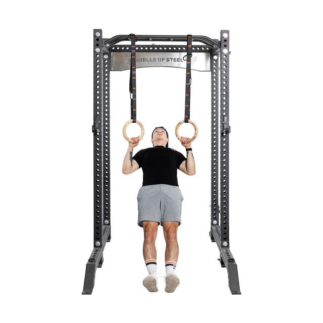 A person in a black shirt and gray shorts demonstrates strength and balance by performing a core workout on Bells of Steel Gymnastics Rings attached to a sturdy metal frame, keeping their feet off the ground to showcase the effectiveness of bodyweight training.