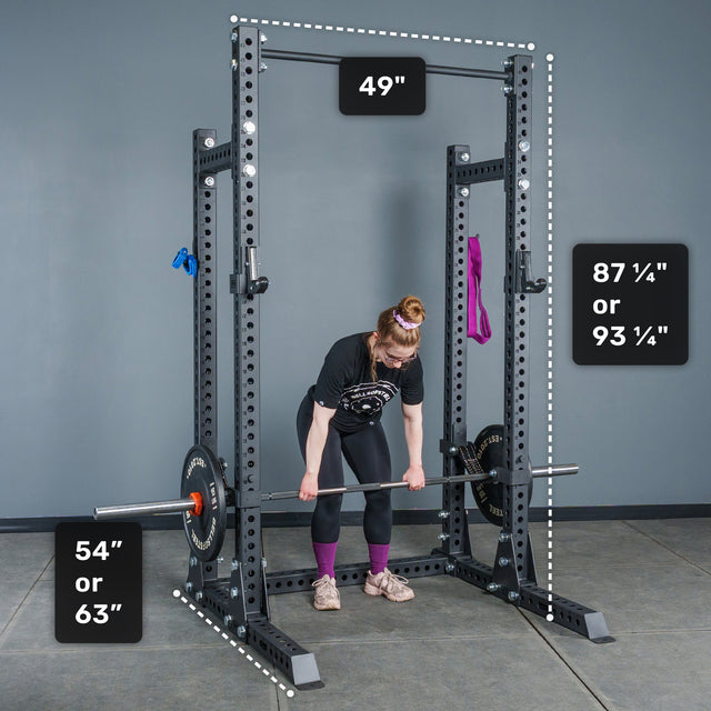 A person lifts a barbell inside the Bells of Steel Manticore Half Rack Builder (3" x 3", 1" Holes), showcasing an ideal home gym setup. The rack features robust 3x3 uprights, with dimensions of 49" width, either 54" or 63" depth, and a choice of 87¼" or 93¼" height. Resistance bands are securely attached to the rack.
