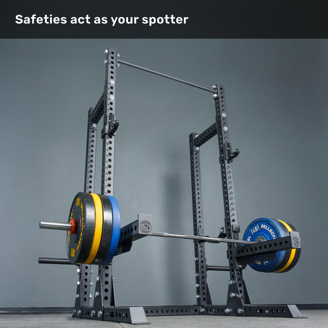 A loaded barbell rests on safeties within the Bells of Steel Manticore Half Rack - Prebuilt (3" x 3", 1" Holes). The text above reads, "Safeties act as your spotter." The background features a plain gray wall and floor—an ideal setup for any home gym.