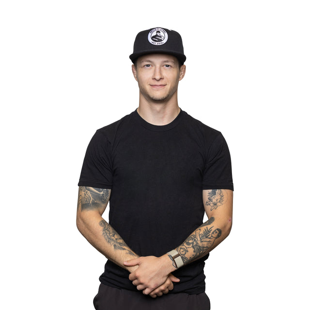 A man wearing a black t-shirt and a Bells of Steel Snap Back Hat with a round logo stands against a white background, smiling with his hands clasped in front. Tattoos are visible on both his arms.