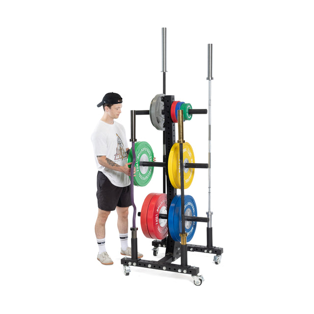 A person in a cap, t-shirt, shorts, and sneakers organizes weight plates on the Bells of Steel Hydra Bumper Plate Tree Builder, a modular storage unit with bars and wheels for easy mobility.