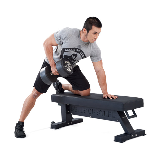 A man in athletic wear uses the Bells of Steel Hero Heavy-Duty Weight Bench to perform a single-arm dumbbell row, supporting himself with one knee and hand, set against a white background.