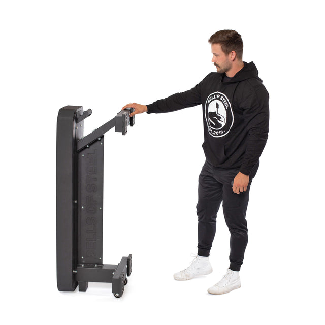 A man in a black hoodie and white sneakers stands beside a folded Bells of Steel Hero Heavy-Duty Weight Bench, holding its frame upright against a white background.
