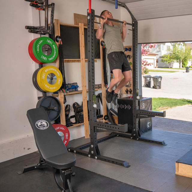 A man uses the Bells of Steel Home Gym Builder squat rack for pull-ups in his home gym. Weight plates, barbells, and other equipment are organized on shelves, while sunlight enters through the open garage door.