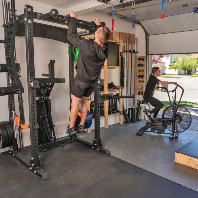 A man does pull-ups on a Bells of Steel Home Gym Builder rack while a woman rides an exercise bike in a well-equipped garage gym with weights, a barbell, and plates.