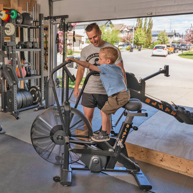 A smiling man supervises a boy riding an air bike in a well-equipped garage gym featuring the Bells of Steel Home Gym Builder, with power racks and weight plates, sunlight streaming through the open door to a sunny street.
