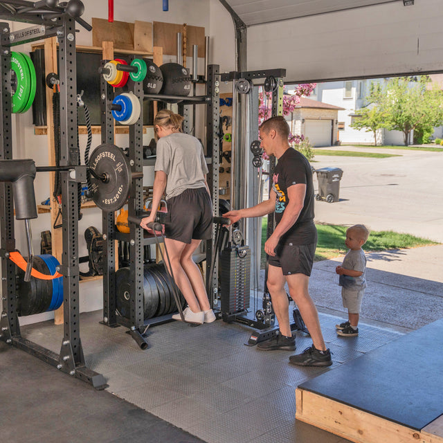 A woman uses the Bells of Steel Home Gym Builder to do triceps dips in her garage gym near a power rack, as a man supports her and a toddler watches. Sunlight pours in through the open garage door.