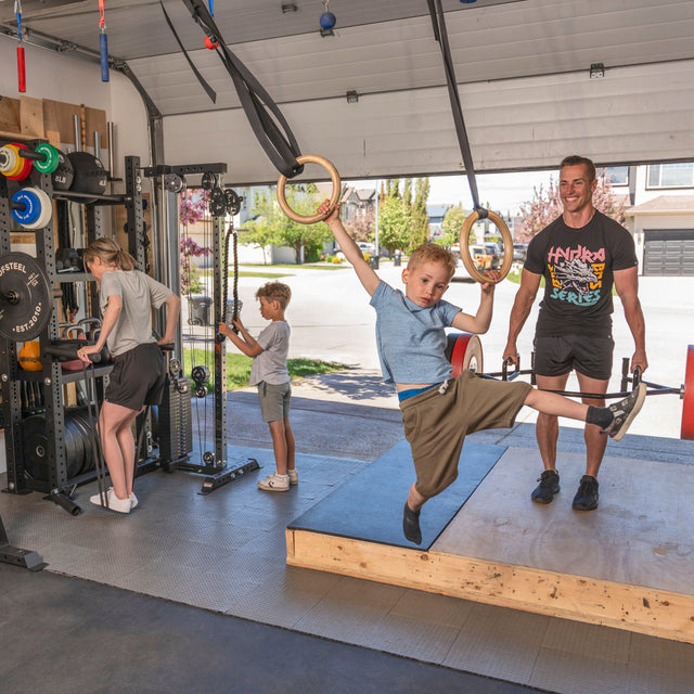 A family enjoys their Bells of Steel Home Gym Builder in the garage: one boy swings on gymnastic rings, another adjusts the equipment by the power rack, a woman lifts weights, and a man smiles with dumbbells as sunlight streams through the open door.