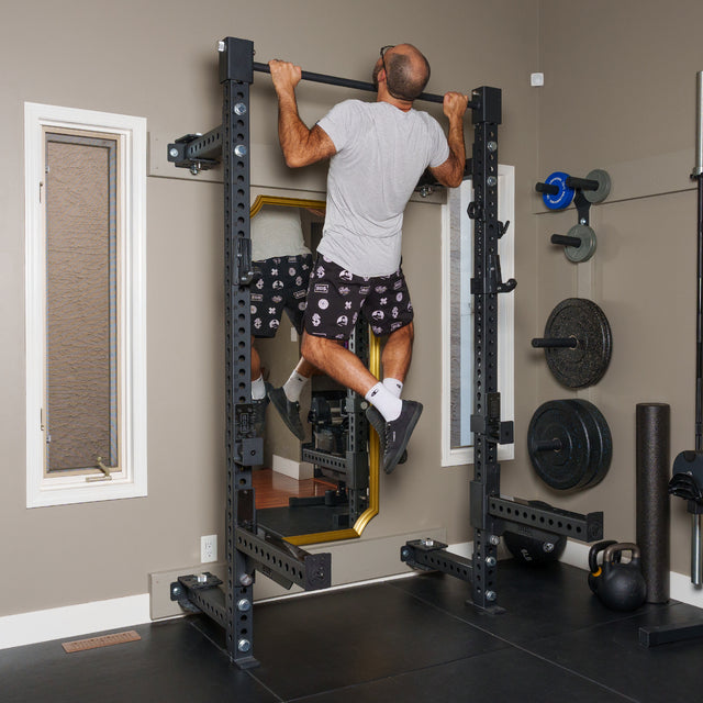 A man in a gray tee and patterned shorts does pull-ups on a Bells of Steel Manticore Folding Half Rack Builder (3" x 3", 1" Holes) in his home gym, with weights, kettlebells, and mirrors seen in the background.