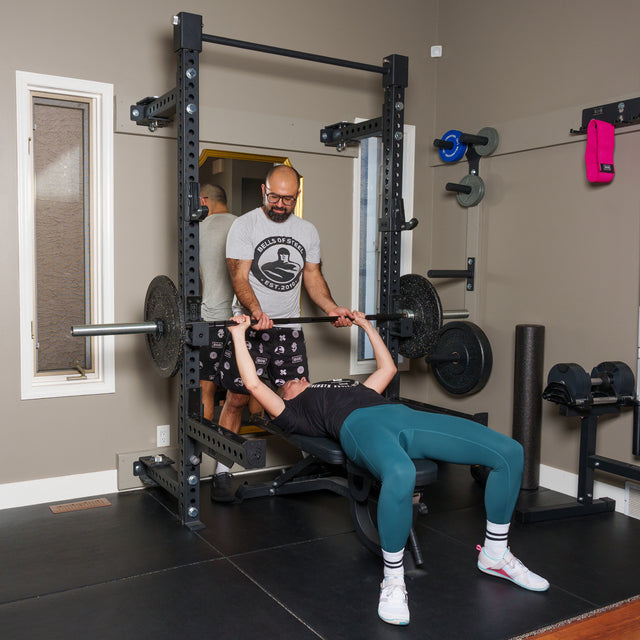In a home gym, someone bench presses on the Bells of Steel Manticore Folding Half Rack Builder (3" x 3", 1" Holes) with teal leggings and white socks, while a spotter in a printed shirt and shorts stands behind.