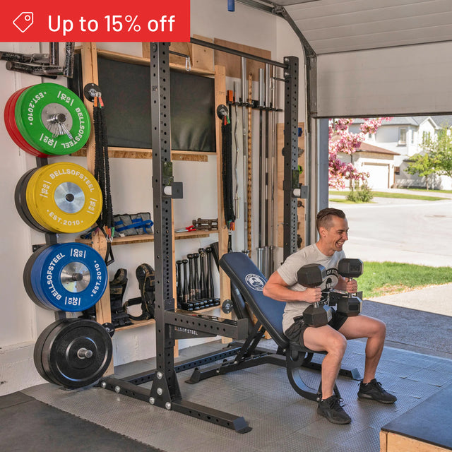 A man smiles on a weight bench in his home gym, holding dumbbells. Bells of Steel Home Gym Builder equipment, including colorful plates and barbells, is organized on the wall. A red banner above reads "Up to 15% off.