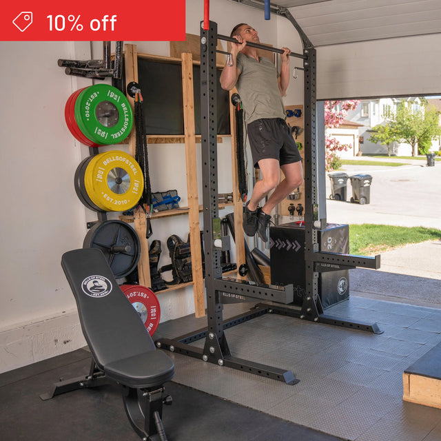 A man uses the Bells of Steel Home Gym Builder for pull-ups in his garage gym, with a weight plate set and bench close by. A red "10% off" banner appears in the corner as sunlight streams through the open door.