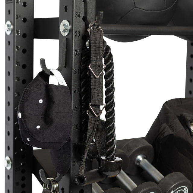 A close-up of Bells of Steel Utility Hooks on a rack with numbered holes holds a black baseball cap, tricep rope attachment, dumbbells, and a medicine ball for an organized workout space.